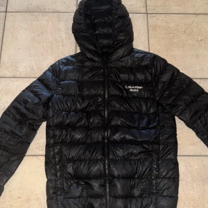 Svart dunjacka från Calvin Klein Jeans - Säljer en svart dunjacka från Calvin Klein Jeans med huva och dragkedja framtill. Jackan har quiltad design och logga på bröstet. Perfekt för kyliga dagar och enkel att matcha med olika outfits.