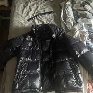 Svart pufferjacka från Polo Ralph Lauren - Säljer en svart pufferjacka från Polo Ralph Lauren med dragkedja och klassisk logga på bröstet. Jackan har hög krage och två sidofickor med dragkedja. Perfekt för kyliga dagar och riktigt snygg till streetwear.