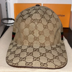 Beige keps från Gucci med GG-mönster - Säljer en beige keps från Gucci med klassiskt GG-monogram över hela kepsen. Kepsen har grönt och rött band på sidan och böjd skärm. Perfekt accessoar för en trendig look.