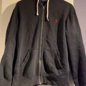 Svart hoodiejacka från Polo Ralph Lauren - Svart hoodiejacka med dragkedja och vit snörning i huvan. Klassisk röd Polo-logga broderad på bröstet. Två fickor framtill och mjukt material som passar perfekt till vardags.