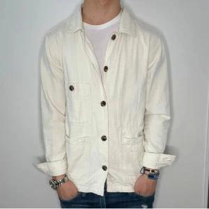 Vit overshirt med knappar - Stilren vit overshirt med stora bruna knappar och tre framfickor. Jackan har en avslappnad passform och är tillverkad i ett bomullsliknande material. Perfekt att bära över en t-shirt för en enkel och snygg look.