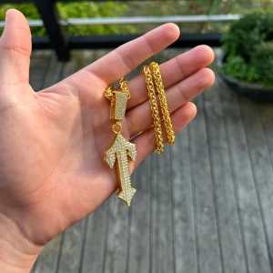 Guldfärgat halsband med glittrig pil-hänge - Snyggt halsband i guldfärg med ett stort pilformat hänge täckt av glittrande stenar. Kedjan är tvinnad och ger ett exklusivt intryck. Perfekt statement piece för dig som vill sticka ut.