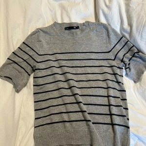 Grå randig tröja från 157 - Grå tröja med svarta horisontella ränder från 157. Tröjan har korta ärmar och rund halsringning. Perfekt för en avslappnad stil och passar bra till jeans eller kjol.