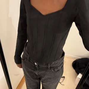 Svart långärmad korsettopp - Snygg svart långärmad korsettopp med markerade sömmar och figurnära passform. Toppen har en djup, fyrkantig urringning och är perfekt för att skapa en trendig look. Passar till både jeans och kjol.