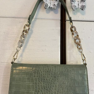 Grön axelväska med kedjedetalj - Snygg grön axelväska från Åhléns med croco-mönstrad yta och bred plastkedja som axelrem. Väskan har ett stilrent och modernt utseende, perfekt för att lyfta din outfit. Flera fack inuti för enkel organisering.