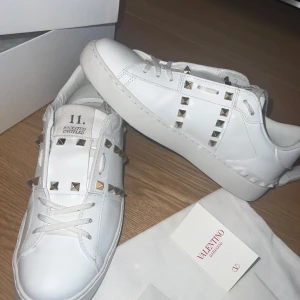 Vita Valentino Rockstud sneakers - Säljer ett par vita Valentino Rockstud sneakers i skinn med ikoniska nitar i silver längs sidorna och över vristen. Klassisk låg modell med rund tå och vita snören. Kommer med originalpåse och extra skosnören. Priset kan diskuteras i snabb affär.