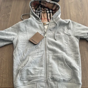 Grå hoodie från Burberry - Snygg grå hoodie från Burberry med klassiskt rutigt foder i huvan och dragkedja framtill. Tröjan har två fickor och justerbar huva med snören. Aldrig använd och väldigt bra skick, passar även S. Priset kan diskuteras 