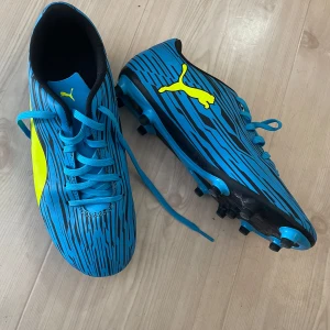 Blå fotbollsskor från Puma - Säljer ett par blåa fotbollsskor från Puma med svart och gult mönster. Skorna har snörning och dobbar för spel på gräs. Perfekta för dig som vill sticka ut på planen! Strl 25,5cm 