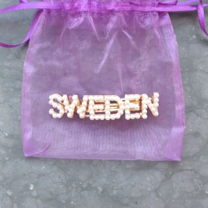 Hårspänne med pärlor och texten SWEDEN💫 - Säljer ett guldigt hårspänne dekorerat med vita pärlor som bildar ordet SWEDEN. Perfekt accessoar för att visa upp din stil och kärlek till Sverige. Kommer i en lila organzapåse.