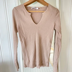 Beige ribbad långärmad topp  - Säljer en beige ribbad långärmad topp från NA-KD med v-ringning och liten slits vid halsen. Perfekt basplagg med smickrande passform och mjukt material.