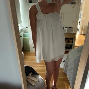 Vit kortklänning med blomdetalj på axeln - Säljer en söt vit kortklänning med breda axelband och en stor tygblomma på ena axeln. Klänningen har en luftig och lös passform. Märke okänd då lappen är bortklippt. 