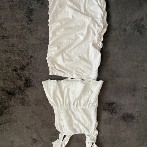 Vit ärmlös klänning med volangdetaljer - Söt vit klänning med broderade detaljer och volanger upptill. Axelbandsmodell med knytband på axlarna och lätt rynkad kjol. Perfekt för varma dagar och somriga tillfällen.