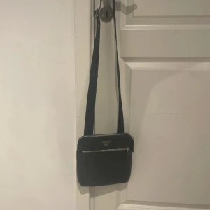 Svart axelväska från Emporio Armani - Snygg svart axelväska från Emporio Armani med silverfärgad dragkedja och logga framtill. Väskan har justerbar rem och ett huvudfack samt ytterfack med dragkedja. Perfekt för att bära det viktigaste. Väskan säljs för 1500 kr i affär just nu 