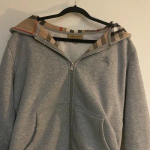 Grå hoodie med dragkedja från Burberry - Säljer en grå hoodie från Burberry med dragkedja och klassiskt rutigt foder i huvan. Tröjan har broderad logga på bröstet och två fickor framtill. Perfekt för dig som gillar stilrena och exklusiva plagg. SAKNAR TYVÄRR SNÖREN TILL LUVAN.