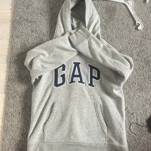 Grå hoodie från GAP Kids - Säljer en klassisk grå hoodie från GAP Kids i storlek L. Tröjan har huva, magficka och stor GAP-logga framtill i mörkblått. Perfekt för en avslappnad stil och enkel att matcha med det mesta.