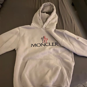 Ljusgrå hoodie från Moncler - Säljer en stilren ljusgrå hoodie från Moncler med broderad logga i blått och rött framtill samt patch på ärmen. Tröjan har huva med dragsko och en stor magficka. Perfekt för dig som gillar exklusiva streetwear-plagg.