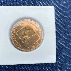 Lokalmynt för Eskilstuna 20 kr - Säljer ett lokalmynt för Eskilstuna med valören 20 kr. Myntet är i kopparfärg och har motiv av en smed samt texten 'LOKALMYNT FÖR ESKILSTUNA'. På baksidan står det 'SKOGHALL' och årtalen 1969-1989 med en byggnad avbildad.