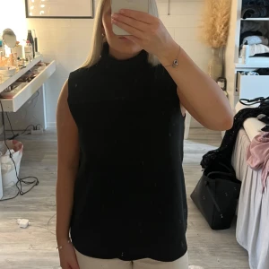 Svart ärmlös polotopp - Säljer en svart ärmlös polotopp med hög hals. Enkel och stilren design som passar till många olika outfits. Perfekt för dig som gillar minimalistisk stil.