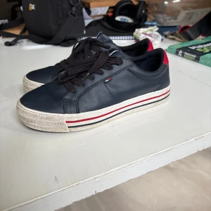 Mörkblå sneakers från Tommy Hilfiger - Säljer ett par mörkblå sneakers från Tommy Hilfiger med svarta skosnören, vit sula med röd och blå rand samt röd detalj på hälen. Klassisk och stilren design med liten logga på sidan.