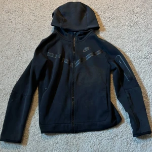 Svart hoodie från Nike -  Storlek 147-158 cm junior. Säljer en svart hoodie från Nike med dragkedja framtill och huva. Jackan har diskreta svarta detaljer över bröstet hoodien har en ficka med dragkedja på ena ärmen. Perfekt för en sportig och stilren look. Några defekter. Skick 5/10