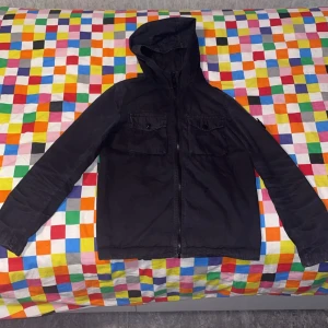 Svart jacka med huva från Stone Island - Säljer en svart jacka från Stone Island med huva och dragkedja. Jackan har två bröstfickor med lock och klassisk Stone Island-logga på ärmen. Perfekt för dig som gillar streetwear och vill ha en stilren look.