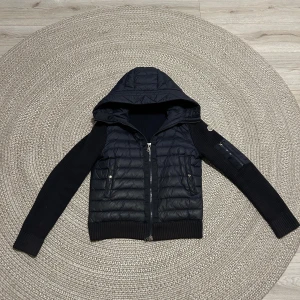 moncler cardigan - Svart pufferjacka med stickade ärmar och huva. Jackan har dragkedja framtill, två fickor med dragkedja och ribbade muddar. Perfekt för kyliga dagar när du vill ha både värme och stil.
