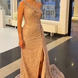 Champagnefärgad glittrig långklänning med slits - Säljer en elegant champagnefärgad långklänning med glittrigt tyg och en axel. Klänningen har en hög slits framtill och ett långt släp, perfekt för bal eller fest. Materialet är glittrigt och ger en lyxig känsla. St 36 men insydd till 34 går att sprätta upp, finns långarm man kan sy till igen på klänningen