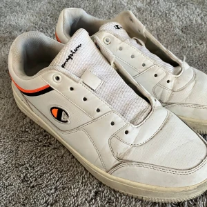 Vita sneakers från Champion med orange detalj - Säljer ett par vita sneakers från Champion med svart och orange rand längs sidan och broderad Champion-logga. Tyvärr saknas skosnören därav priset.