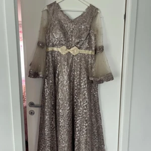 Champagnefärgad långklänning med spets och pärlor - Elegant långklänning i champagnefärg med glittrigt spetsmönster, pärlprydda transparenta ärmar och vacker rosettdetalj i midjan. Klänningen har snörning i ryggen och utsvängda ärmar för en extra lyxig känsla.