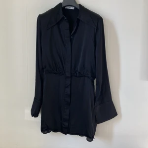 Svart skjortklänning från MNG - Stilren svart skjortklänning från MNG med markerad midja och klassisk krage. Klänningen har långa ärmar med breda manschetter och knäppning framtill. Perfekt för dig som gillar en enkel men elegant look.