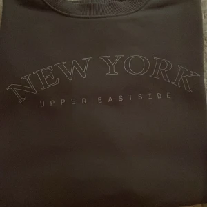 Mörkgrå sweatshirt med New York-tryck - Säljer en mörkgrå sweatshirt med texten 'NEW YORK UPPER EASTSIDE' tryckt på bröstet. Tröjan har rund halsringning och långa ärmar. Perfekt för en avslappnad och trendig stil.