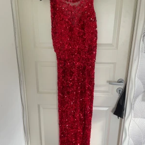 Röd paljettklänning med slits - Säljer en lång, röd klänning täckt av paljetter med v-ringning och transparent mesh upptill. Klänningen har en snygg slits på sidan och är ärmlös, perfekt för bal eller fest. Båtrygg. Använd 1 gång