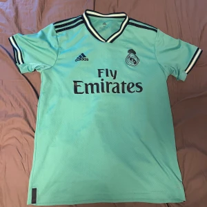 Real Madrid fotbollströja Adidas - Snygg ljusgrön Real Madrid fotbollströja från Adidas med svarta detaljer och klubbmärke på bröstet. Tröjan har korta ärmar, svart Adidas-logga och Fly Emirates-tryck på framsidan. Perfekt för dig som älskar fotboll och vill sticka ut på planen.