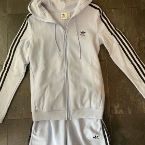 Adidas sett  - Säljer ett mjukissett från Adidas med klassiska svarta ränder längs sidorna och resår vid bensluten.  Dekorativa knappar vid armar och vid benslut. Perfekta för en avslappnad stil.