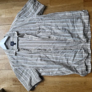 Beige och vit randig skjorta till sommaren  - Kortärmad skjorta i beige och vit färg med tunna ränder och en bröstficka. Perfekt för varma dagar och enkel att matcha med jeans eller shorts. Storleken är small, så den passar ca 170-180. Priset är inte satt i sten, så kontakta gärna! 