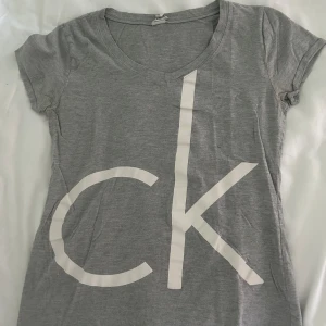 Calvin Klein T-shirt - Grå t-shirt från Calvin Klein med stor vit CK-logga framtill. T-shirten har en lagom stor v-ringad hals. Den är i storlek M men passar även S