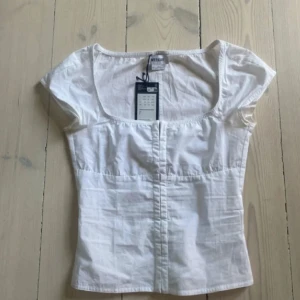 Vit topp med fyrkantig ringning från Weekday - Säljer en vit topp från Weekday med korta ärmar och fyrkantig ringning. Toppen har en figurnära passform och är tillverkad i bomullsmaterial. Perfekt att styla till jeans eller kjol. 