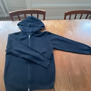 Mörkblå stickad hoodie med dragkedja - Säljer en mörkblå stickad hoodie med dragkedja framtill. Tröjan har långa ärmar, huva och ribbade muddar vid ärmslut och nederkant. Perfekt för kyliga dagar och enkel att matcha med olika outfits.