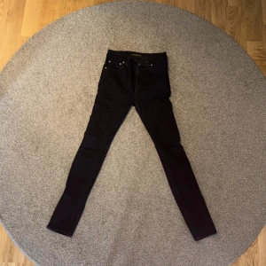 Svarta jeansbyxor från Nudie Jeans - Säljer ett par svarta jeansbyxor från Nudie Jeans, helt oanvända utan prislapp. Klassisk femficksdesign och smal passform. Byxorna har hög midja och är tillverkade i ett stretchigt bomullsmaterial. Perfekta för dig som gillar en stilren och enkel look.