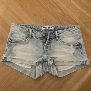 Lågmidjade jeansshorts - Snygga ljusblå lågmidjade jeansshorts med slitna detaljer och fransig kant. Modellen har tre knappar framtill, klassiska fickor och låg midja. Perfekta för varma dagar och en avslappnad stil.