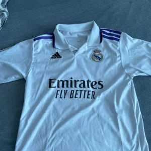 Real Madrid fotbollströja Modrić 10 - Säljer en vit Real Madrid fotbollströja från Adidas med Modrić och nummer 10 på ryggen. Tröjan har korta ärmar, klubbmärke på bröstet och sponsortryck framtill. Klassisk design med blå detaljer på axlarna.