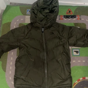 Mörkgrön pufferjacka med huva - Säljer en mörkgrön pufferjacka från Nike med huva och dragkedja framtill. Jackan har två fickor på sidorna och är perfekt för kyligare dagar. Enkel och stilren design med diskret logga på bröstet.
