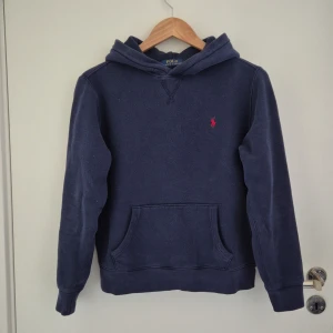 Mörkblå hoodie från Polo Ralph Lauren - Mörkblå hoodie från Ralph Lauren med klassisk känguruficka och huva. Tröjan har en röd broderad logga på bröstet och är tillverkad i mjukt material. Perfekt till sommaren och för en avslappnad stil. Strl 160cm, och den är såklart 100% äkta. INGA SKAMBUD!!!