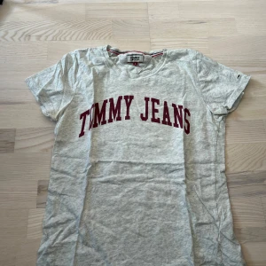Grå t-shirt från Tommy Jeans - Säljer en ljusgrå t-shirt från Tommy Jeans med röd text framtill. Klassisk rund halsringning och korta ärmar. Perfekt för en avslappnad och stilren look.