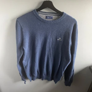 Blå stickad tröja från GANT - Säljer en klassisk blå stickad tröja från GANT med rund halsringning och diskret logga broderad på bröstet. Tröjan har långa ärmar och ribbade muddar vid ärmslut och nederkant. Perfekt för lager-på-lager-stil.