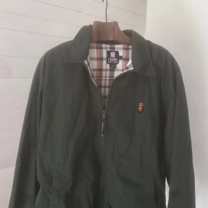 Mörkgrön jacka från Chaps Ralph Lauren - Säljer en mörkgrön jacka från Chaps Ralph Lauren med klassiskt rutigt foder. Jackan har dragkedja framtill, två fickor och en liten broderad logga på bröstet. Materialet är en blandning av polyester och bomull.