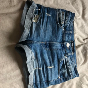 Blå jeansshorts  - Säljer ett par blå jeansshorts med slitna detaljer och upprullade benslut. Klassisk femficksmodell med knapp och dragkedja framtill. Perfekta för varma dagar.