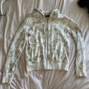 Vit zip-up med tryck  - Vit hoodie med dragkedja och huva, täckt av ett unikt mönster med tecknade änglar i beige/brunt. Jackan har långa ärmar och ribbade muddar. Perfekt för dig som vill sticka ut med en cool och annorlunda stil.