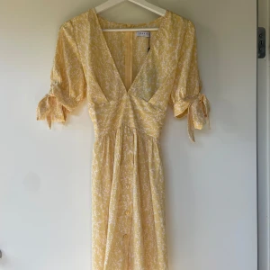 Gul blommig omlottklänning från Loavies - Supersöt gul klänning från Loavies med vitt blommönster, v-ringning och knytband i midjan. Klänningen har korta ärmar med knyt och omlottkänsla framtill. Perfekt för varma dagar och somriga tillfällen!