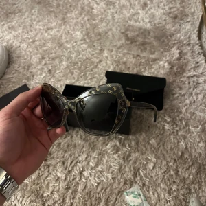 Svarta solglasögon från Dolce & Gabbana - Snygga svarta solglasögon från Dolce & Gabbana med stora, kantiga bågar och diskret mönster i guld längs kanterna. Glasögonen har mörka linser och en exklusiv känsla, perfekt för att lyfta din stil.